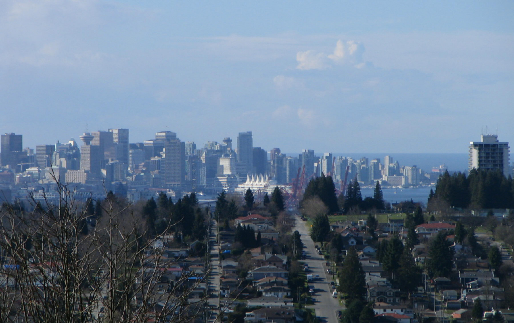 Tree-lined residential streets of single-family homes sloping down toward Burrard Inlet with the Downtown Vancouver skyline beyond, seen from Capitol Hill, Burnaby Heights