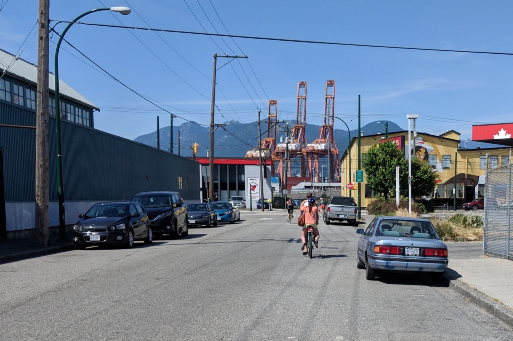 Street sloping toward the waterfront past low industrial buildings and parked cars, with Port of Vancouver container cranes and the North Shore mountains beyond, in Grandview-Woodland, Vancouver