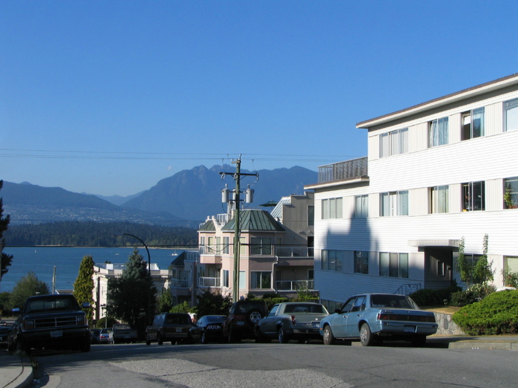 Low-rise apartment buildings and parked cars on a sloping residential street running down toward English Bay, with the North Shore mountains behind, Kitsilano, Vancouver