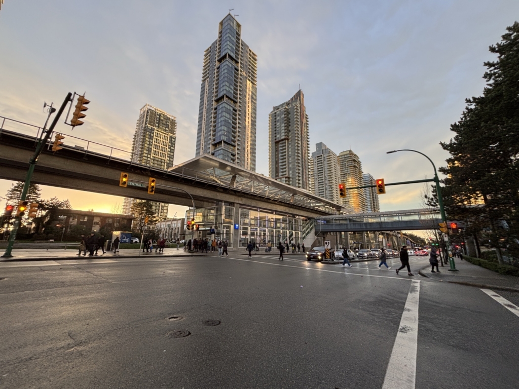 Pedestrians crossing a wet street under the elevated SkyTrain guideway at Central Boulevard, with high-rise condo towers behind, Metrotown, Burnaby