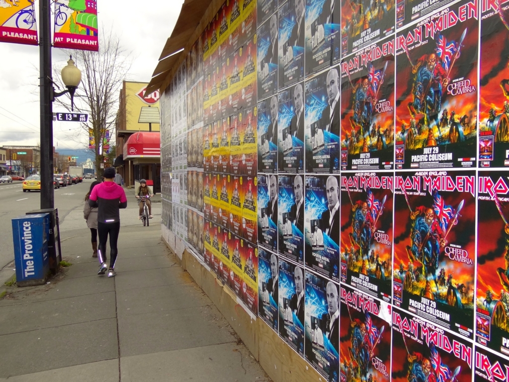 Pedestrians and a cyclist on a commercial street sidewalk past a poster-covered hoarding, under a purple MT PLEASANT banner beside the East 13th Avenue sign, Mount Pleasant, Vancouver