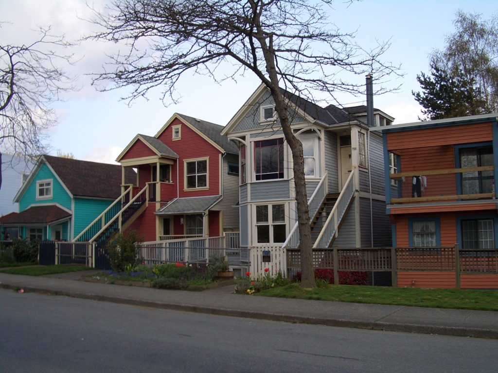 Row of brightly painted heritage wood-frame houses with raised front porches, front gardens and a grass boulevard along a residential street in Strathcona, Vancouver